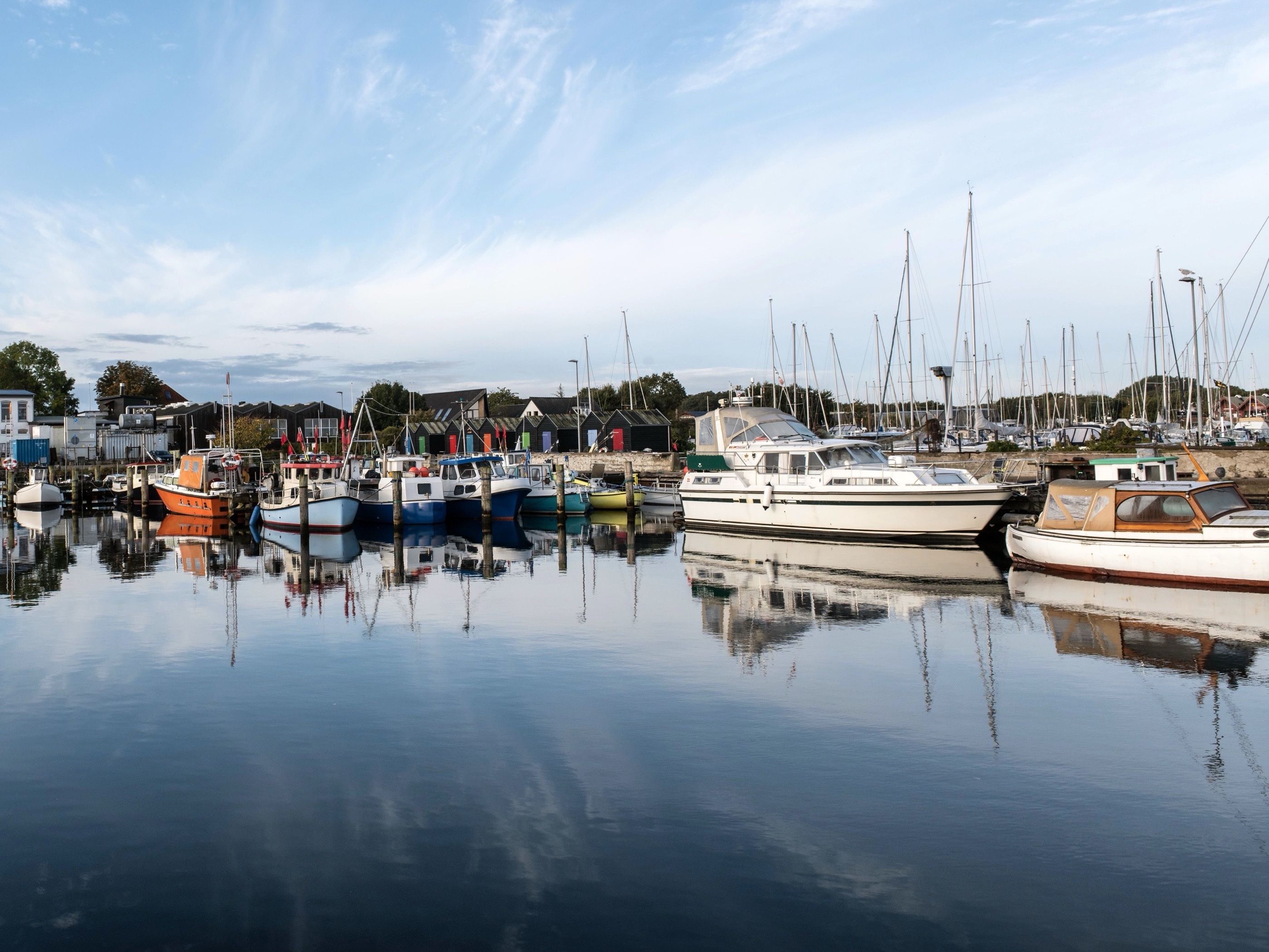 Småbåtshamn i morgonljus. Orange, blå och vita båtar ligger i en liten hamn med master bakom sig, och vattnet är lugnt och speglar dem.