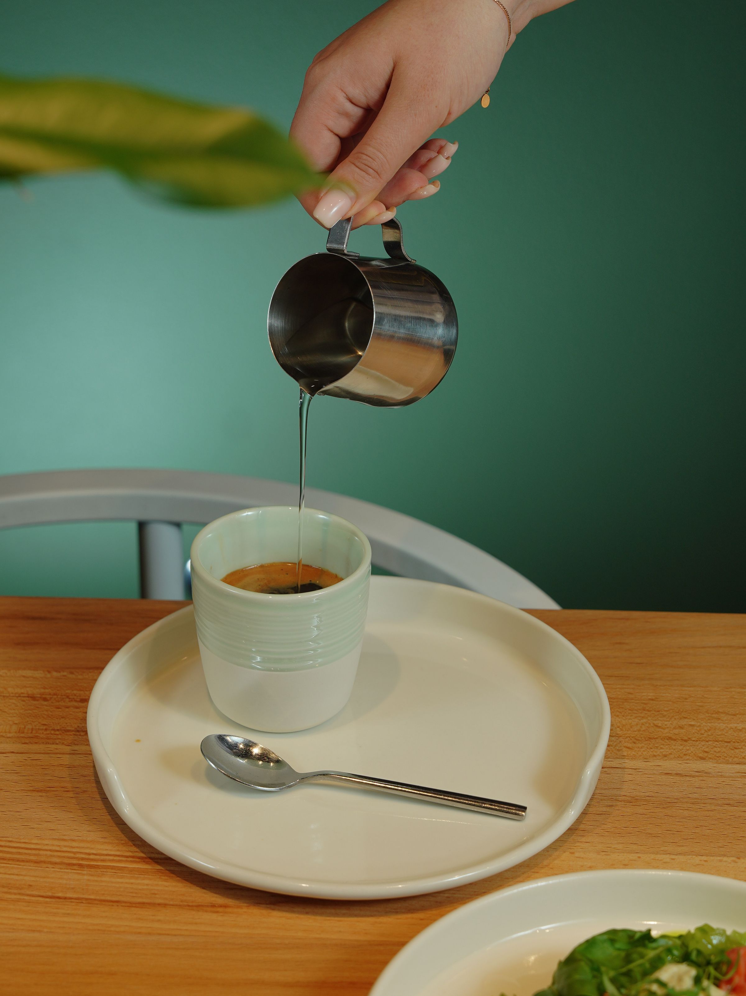 En hand häller från en stålkanna i en espressokopp på ett vitt fat, grön vägg i bakgrunden.